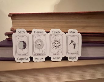 May include: Four white tarot card stickers with black illustrations and text. The cards are labeled "Seth", "Darius", "Caleb", and "Max". The cards are labeled with the names of the major arcana cards: "The Moon", "Strength", "The Sun", and "Temperance". The cards are also labeled with the names of stars: "Capella", "Acrux", "Altair", and "Rigel".