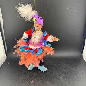 May include: A decorative figurine of a performer in a colorful costume. The figure wears a purple hat with a white feather, a red jacket, and a skirt of orange feathers. Gold trim and blue shoes complete the look.