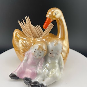 Pode incluir: Um porta-palitos de dentes de porcelana em forma de cisne com duas crianças pequenas sentadas nas suas costas. O cisne é dourado com um bico vermelho e as crianças usam roupas rosa e brancas.