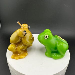 May include: Two frog-shaped candles. One is a golden yellow colour, and the other is bright green. The candles are on a white rotating platform against a black background. The candles have wicks.