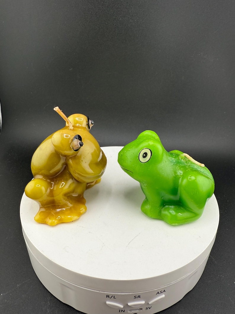 May include: Two frog-shaped candles. One is a golden yellow colour, and the other is bright green. The candles are on a white rotating platform against a black background. The candles have wicks.