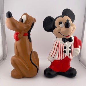Puede incluir: Un conjunto de figuras de cer&aacute;mica con Mickey Mouse y Pluto. Mickey lleva una camisa a rayas rojas y blancas, pantalones rojos y una pajarita negra. Pluto es marr&oacute;n con una pajarita roja.