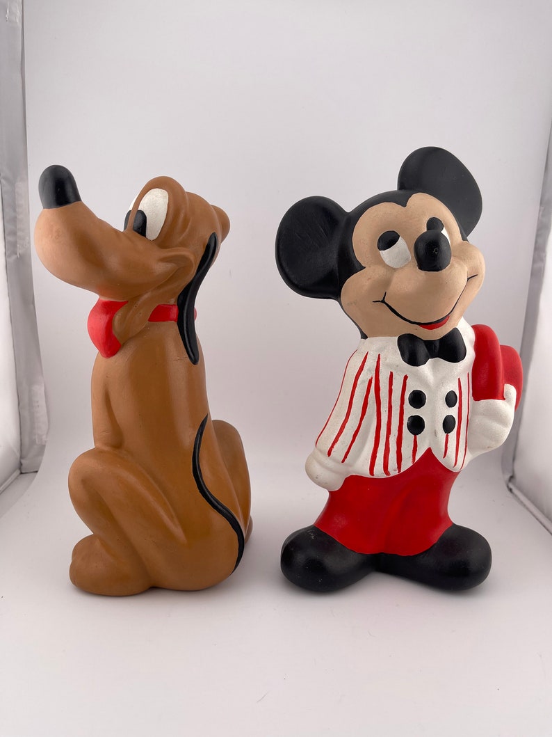 Puede incluir: Un conjunto de figuras de cer&aacute;mica con Mickey Mouse y Pluto. Mickey lleva una camisa a rayas rojas y blancas, pantalones rojos y una pajarita negra. Pluto es marr&oacute;n con una pajarita roja.