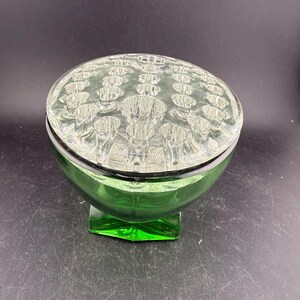 May include: A green glass flower frog with a clear, textured lid. The base is a rounded bowl shape, and the lid has multiple holes for arranging flowers. The frog sits on a square base.