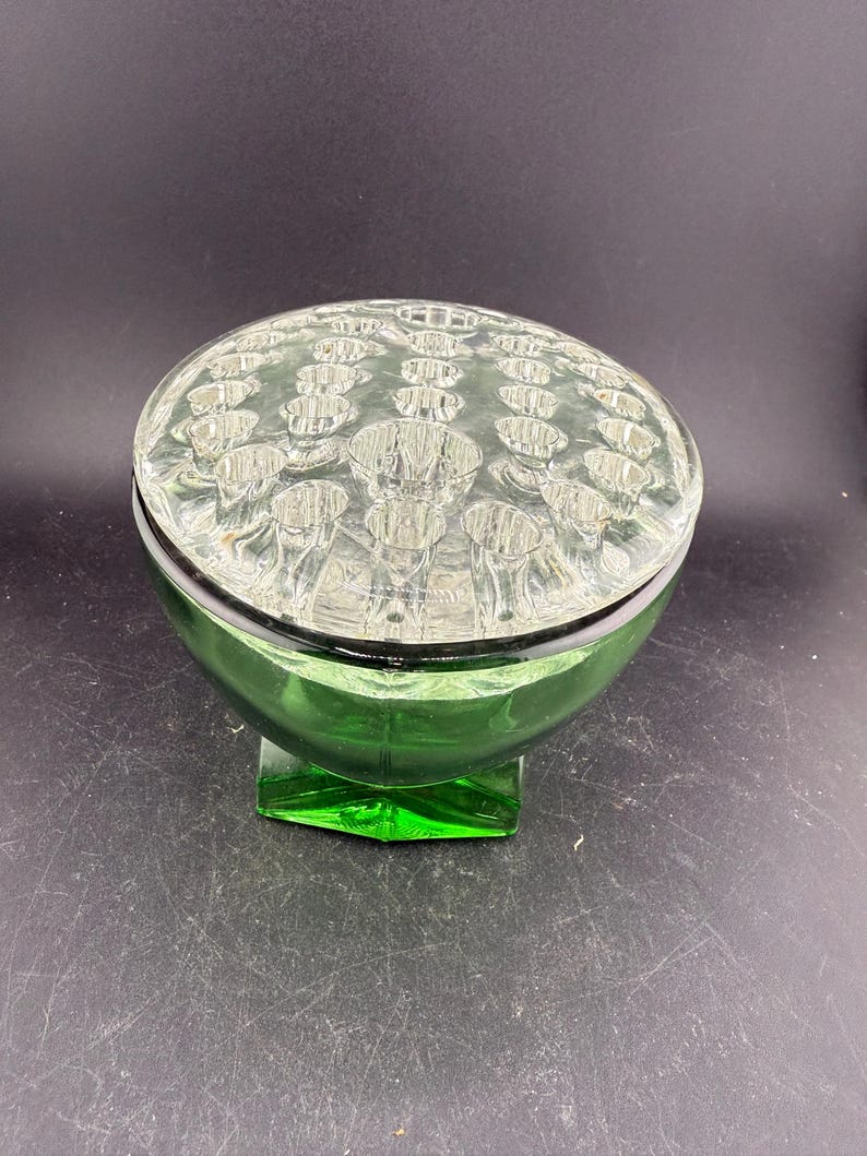 May include: A green glass flower frog with a clear, textured lid. The base is a rounded bowl shape, and the lid has multiple holes for arranging flowers. The frog sits on a square base.