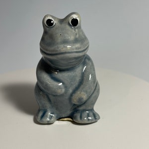 May include: A small, grey ceramic frog figurine with black eyes and white pupils. The frog is sitting upright with its legs tucked in.