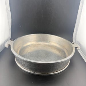 May include: A round, shallow metal pan with a smooth interior and a textured, hammered-style exterior finish. The pan features two integrated, decorative handles on opposite sides, each with a small cutout detail at the top edge.