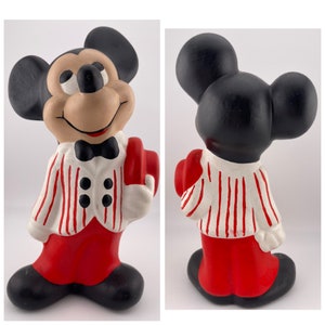 Puede incluir: Una figurita de cer&aacute;mica de Mickey Mouse con una chaqueta a rayas rojas y blancas, pantalones rojos y una pajarita negra. Lleva un sombrero rojo en su mano derecha.