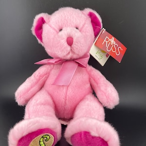 May include: A pink plush teddy bear with dark pink accents on the ears and feet. The bear has a pink bow and a tag that reads "RUSS Aries". The bear's feet have a gold emblem with a green design and the word "ARIES".