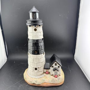 Puede incluir: Una figura de faro de cerámica con un diseño en blanco y negro. El faro tiene una parte superior negra y una pequeña casa adjunta a la base. La base es de color canela y la casa tiene puertas rojas.