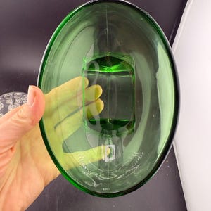 May include: A green glass oval dish with a rectangular base. The transparent dish is held by a hand. The background is dark. The dish is likely for decorative or serving purposes.
