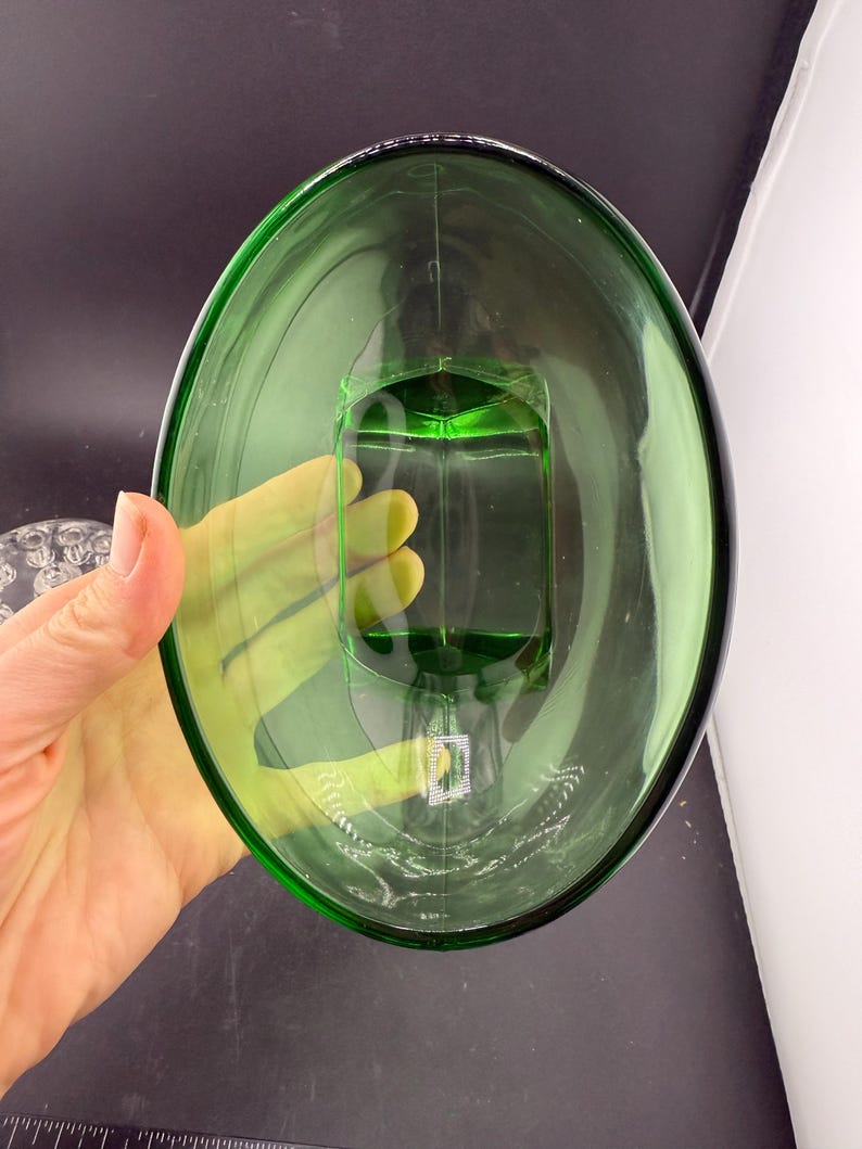 May include: A green glass oval dish with a rectangular base. The transparent dish is held by a hand. The background is dark. The dish is likely for decorative or serving purposes.