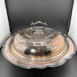 Puede incluir: Una fuente plateada con tapa abovedada y asa decorativa. La fuente tiene un borde festoneado y un monograma en la tapa. La fuente está diseñada para servir alimentos.