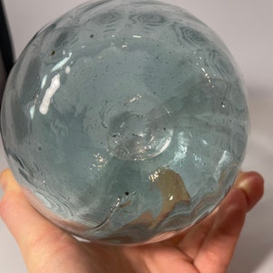 May include: A round, light blue glass float with a textured surface. The float has a small hole at the bottom.