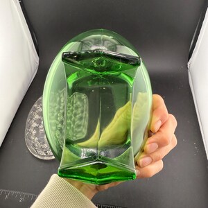 May include: A green glass decanter with a geometric design. The decanter has a square base and a rounded top. A clear glass plate with a floral design is visible in the background.