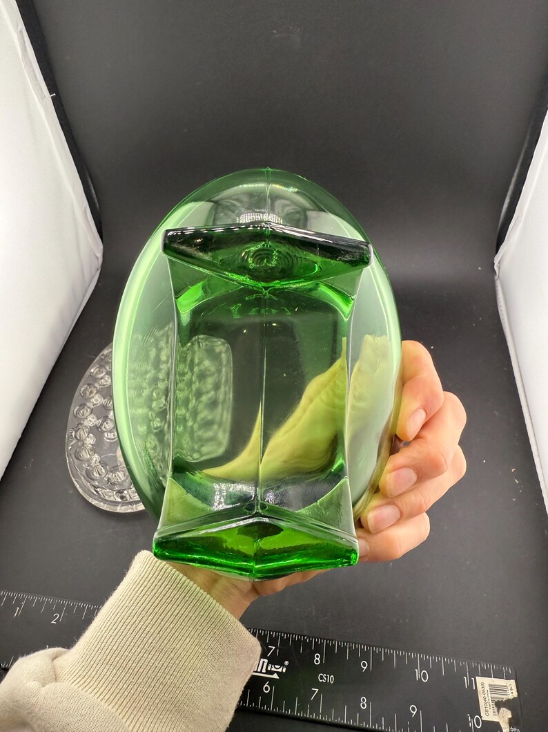 May include: A green glass decanter with a geometric design. The decanter has a square base and a rounded top. A clear glass plate with a floral design is visible in the background.