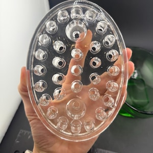 May include: Clear, oval flower frog with numerous holes for arranging stems. The glass frog is held in a hand, with a green glass bowl visible in the background. A ruler is in the background.