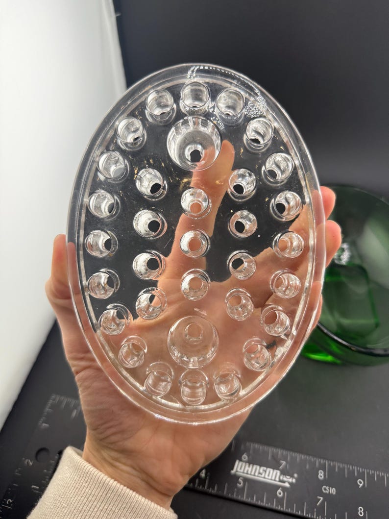 May include: Clear, oval flower frog with numerous holes for arranging stems. The glass frog is held in a hand, with a green glass bowl visible in the background. A ruler is in the background.