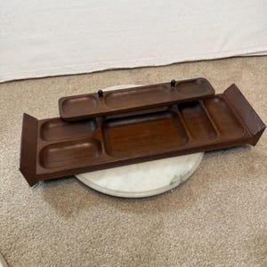 May include: A dark brown wooden serving tray with multiple rectangular compartments and two small black knobs on the top tier. The tray features a tiered design with a smaller, elevated section resting above a larger, wider base with rounded edges.