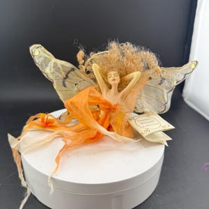 May include: A fairy figurine with orange and cream fabric wings and dress. The fairy has light brown curly hair and arms raised. The wings have a delicate pattern. The figurine is on a white circular base. The wings are adorned with small, sparkling embellishments.