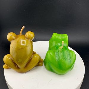 May include: Two frog-shaped candles. One is a muted olive green, the other is a bright green. Both candles have a wick and are sitting on a white surface. The background is black.