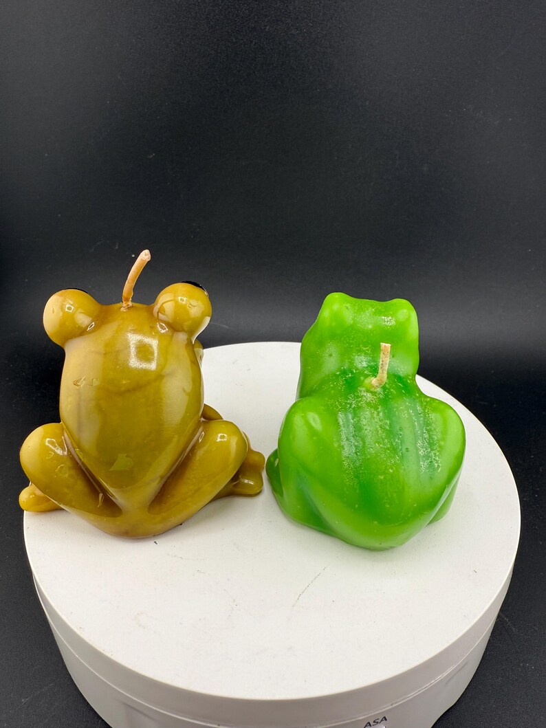 May include: Two frog-shaped candles. One is a muted olive green, the other is a bright green. Both candles have a wick and are sitting on a white surface. The background is black.