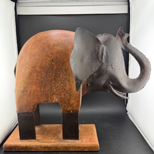 May include: A rustic metal elephant sculpture with a brown body and dark gray ears and trunk. The elephant stands on a rectangular wooden base. The sculpture has a weathered, antique appearance.