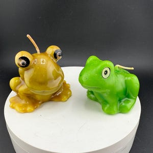 May include: Two frog-shaped candles. One is a golden yellow colour with large, bulging eyes. The other is a bright green frog candle with a more traditional frog appearance. Both candles have wicks.