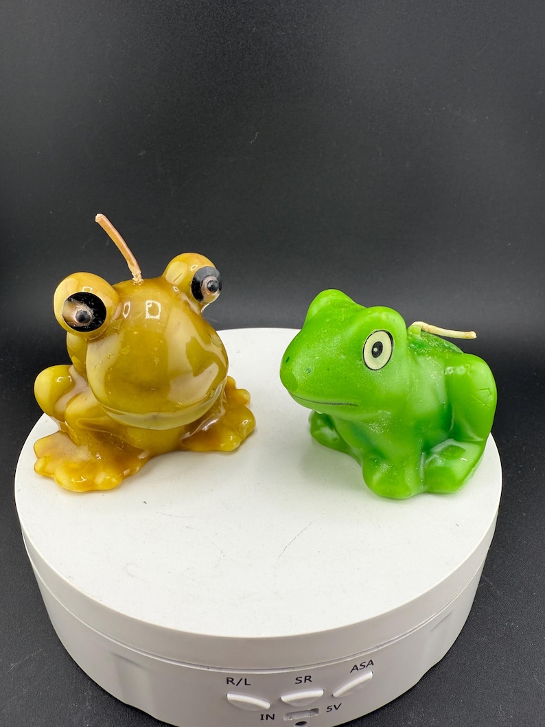 May include: Two frog-shaped candles. One is a golden yellow colour with large, bulging eyes. The other is a bright green frog candle with a more traditional frog appearance. Both candles have wicks.