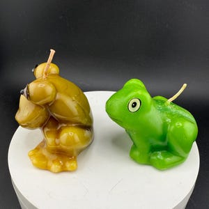 May include: Two frog-shaped candles. One is a golden yellow colour, and the other is bright green. Both candles have a wick extending from the top. The candles are sitting on a white, circular surface against a black background.