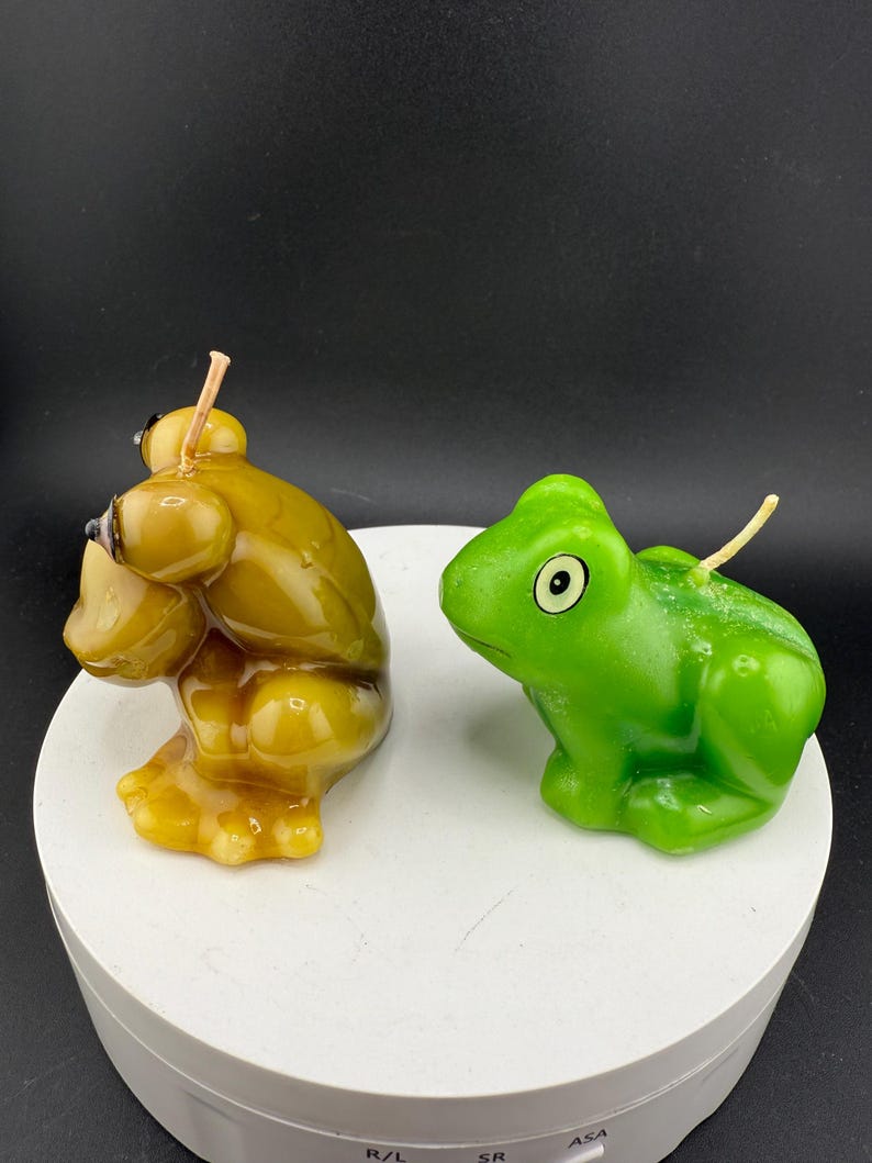 May include: Two frog-shaped candles. One is a golden yellow colour, and the other is bright green. Both candles have a wick extending from the top. The candles are sitting on a white, circular surface against a black background.