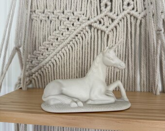 May include: A white ceramic unicorn figurine in a resting pose. The mythical creature has a single horn and is lying on a small, oval base. The background features a macrame wall hanging and a wooden shelf.