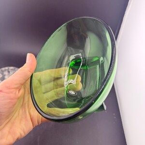 May include: A clear green glass bowl with a rounded shape and a black rim. The bowl is held by a hand, and the interior reflects light. The bowl is empty and appears to be made of thick glass.