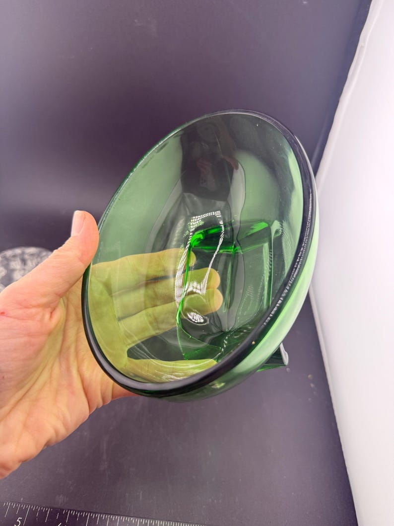 May include: A clear green glass bowl with a rounded shape and a black rim. The bowl is held by a hand, and the interior reflects light. The bowl is empty and appears to be made of thick glass.