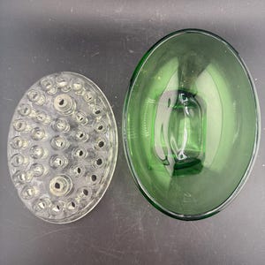 May include: Two glass flower arranging tools. A clear, oval-shaped flower frog with multiple holes sits next to a green, oval-shaped glass bowl. The bowl has a rectangular base and is approximately 25 cm long.