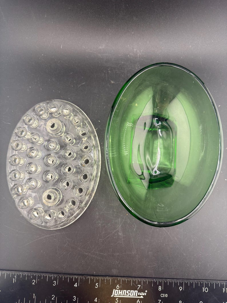 May include: Two glass flower arranging tools. A clear, oval-shaped flower frog with multiple holes sits next to a green, oval-shaped glass bowl. The bowl has a rectangular base and is approximately 25 cm long.