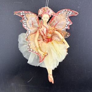 May include: A fairy ornament with butterfly wings. The fairy is wearing a yellow and cream-coloured dress with a tulle skirt. The wings are a mix of cream, orange, and red with glitter accents. The fairy has a delicate face and is wearing gold bracelets.