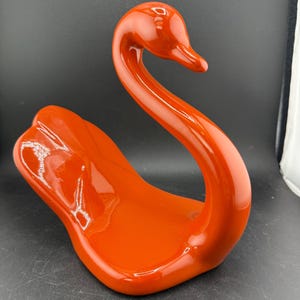Puede incluir: Una figura de cisne de cerámica naranja brillante. El cisne tiene un cuello curvo y un diseño elegante y moderno. La figura es un artículo decorativo, probablemente para una estantería o una mesa.