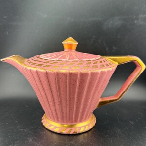 Puede incluir: Una tetera de cerámica rosa con detalles dorados. La tetera tiene un diseño acanalado y una tapa con una punta puntiaguda.