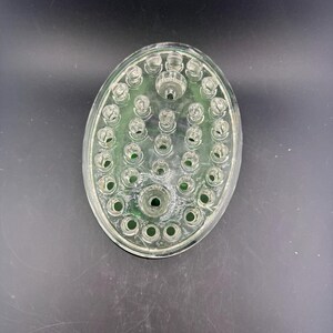 May include: Clear glass oval flower frog with multiple holes for arranging stems. The flower frog has a raised centre and a green tint. The design is for floral arrangements.