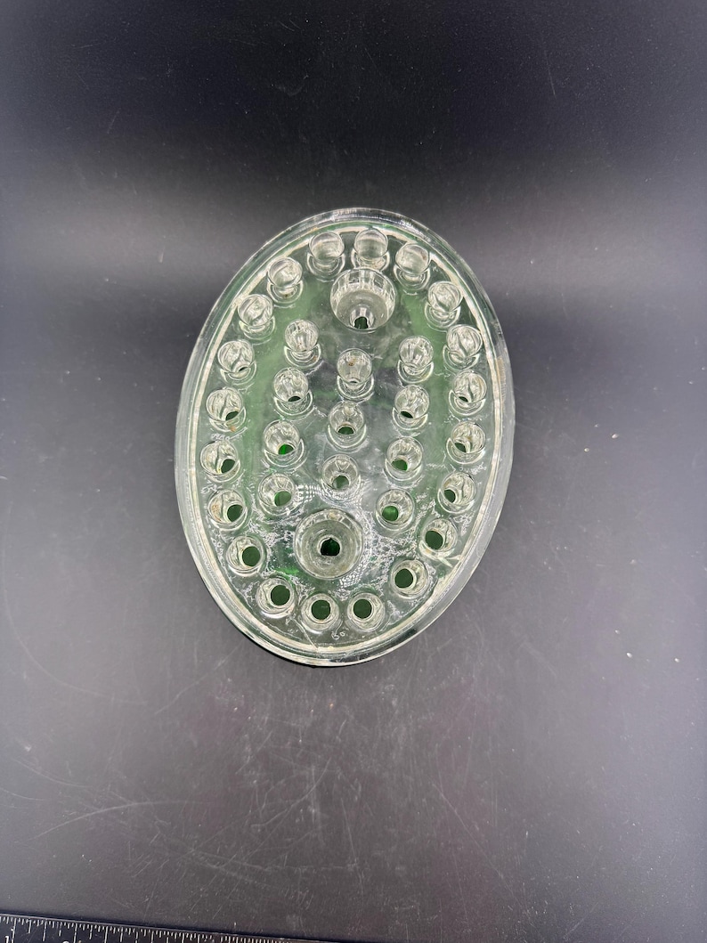May include: Clear glass oval flower frog with multiple holes for arranging stems. The flower frog has a raised centre and a green tint. The design is for floral arrangements.