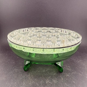 May include: A green glass flower frog with a clear, textured top. The oval bowl rests on three curved feet. The top features a grid pattern of small holes for arranging flowers.