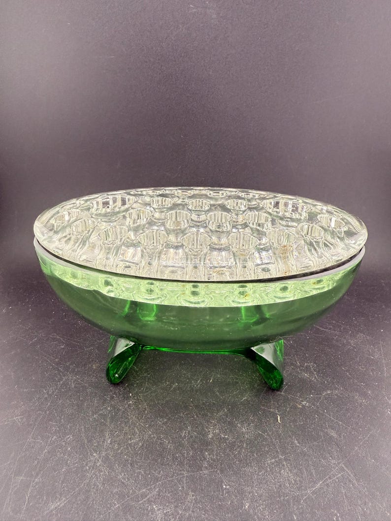 May include: A green glass flower frog with a clear, textured top. The oval bowl rests on three curved feet. The top features a grid pattern of small holes for arranging flowers.
