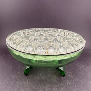 May include: A green glass flower frog with a clear, grid-patterned top. The base is a vibrant green, supported by three curved feet. This vintage piece is designed to hold flowers in place.