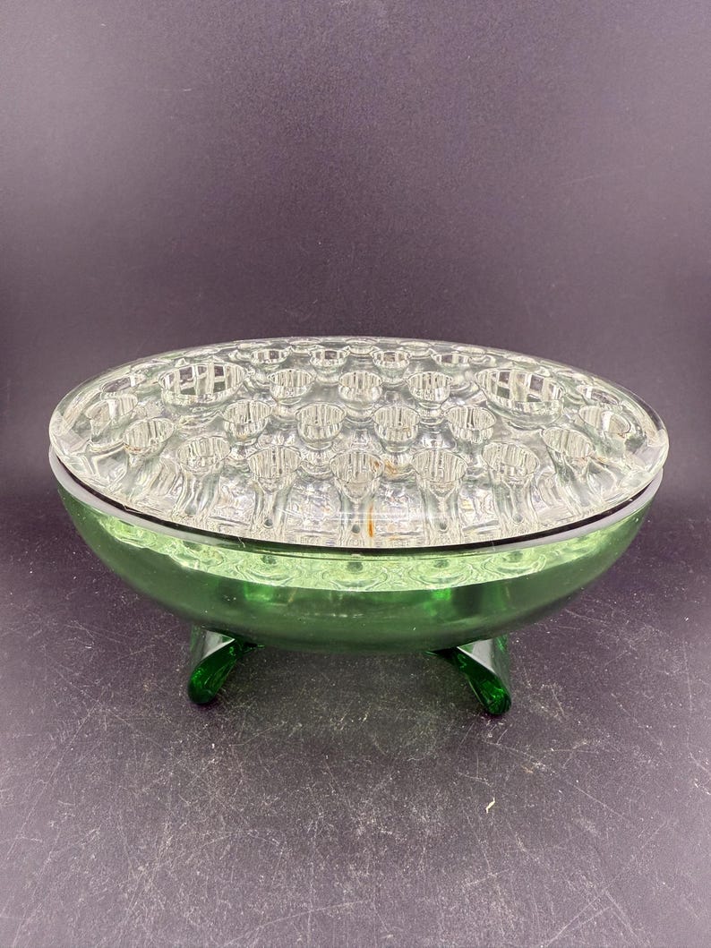 May include: A green glass flower frog with a clear, grid-patterned top. The base is a vibrant green, supported by three curved feet. This vintage piece is designed to hold flowers in place.