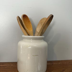 May include: A white ceramic jar with the text "LAB-Lagnu" embossed on the front, holding several wooden spoons. The spoons are various shades of brown and have a well-worn appearance. The jar sits on a wooden shelf.