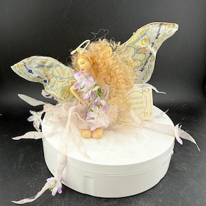 May include: A handcrafted fairy figurine with a halo, holding purple flowers. The fairy has curly blonde hair and delicate, patterned wings. The figurine is wearing a light purple dress and is on a white circular base. A tag reads "Florence Jaranuk Collection."