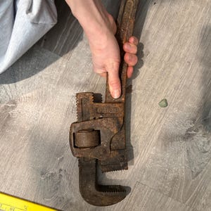 May include: A rusty, vintage pipe wrench. The wrench is a dark brown color, showing signs of age and wear. The jaws are open, and the handle is textured for grip. The tool is designed for gripping and turning pipes.