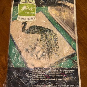 Puede incluir: Un primer plano de un paquete envuelto en plástico que contiene un kit de manualidades para una alfombra de pavo real. El kit incluye hilo de varios colores, una aguja de punzón e instrucciones. El tamaño de la alfombra terminada es de 61 cm por 91 cm.