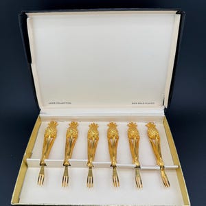May include: Set of six gold-plated pineapple-shaped cocktail forks in a black box with a white interior. The box has the text "JANS COLLECTION" and "24 K GOLD PLATED" printed on it.